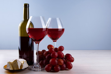 Low angle perspective with a two glass filled with dark red wine and red grape and bootle with white cheese and a thin leg on a wooden table with a light gray background with copy space for your text