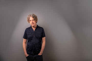 Studio shot of an attractive forty something man in a blue shirt, standing in front of a gray wall.