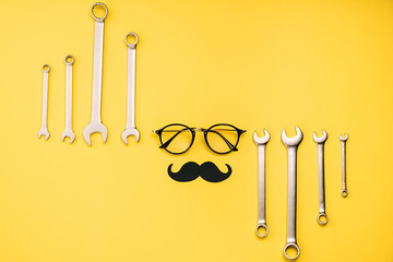 Creative flat lay composition with transparent glasses, stylish black paper photo booth props moustaches and combination wrenches on yellow background copy space. Fathers day masculinity concept blog