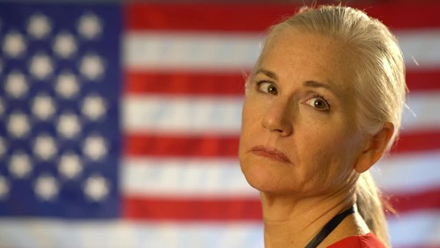Closeup portrait of the back of blonde nurses head as she turns and looks at camera looking concerned and sad with out of focus US flag.