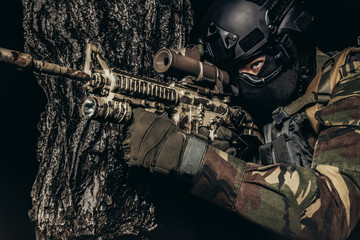 Soldier in uniform sitting and aiming rifle in forest.