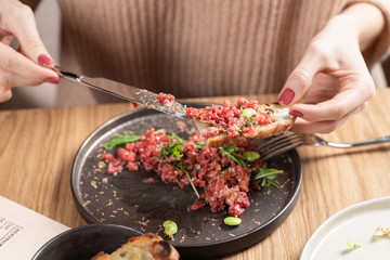 Young woman is eating beef tartare with egg yolk, using knife and fork, mixing up the meal