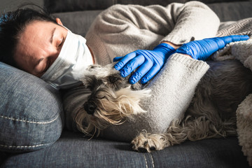 Sick woman with mask and medical gloves hugs a dog lying on the sofa