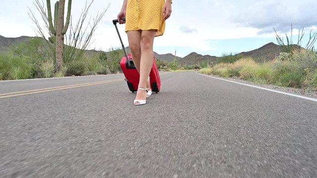 Attractive blond woman pulling her red luggage and walking away from it all on an empty countryside road that leads through thorny cactuses in the Sonoran desert, Arizona