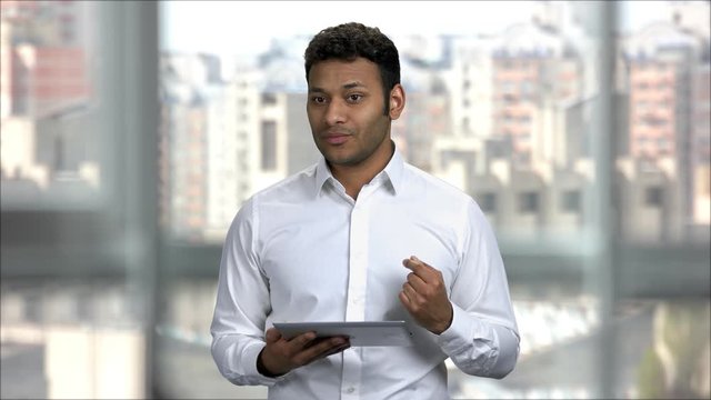 Indian man giving presentation speech in office holding tablet pc. Young asian speaker in white shirt talking and teaching, cityscape view.