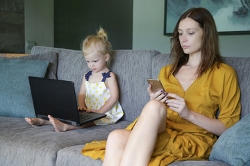 Fototapeta na wymiar Mother and her daughter sitting on sofa in living room spending time on laptop and cellphone, modern family living, technology and internet addiction concept