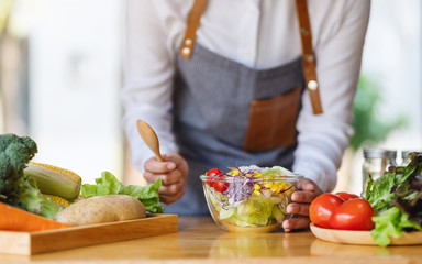 Closeup image of a female chef cooking and holding a bowl of fresh mixed vegetables salad and wooden spoon in kitchen