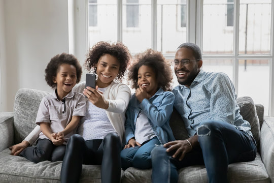 African ethnicity couple with little kids resting on couch having fun holding smart phone using new free cool application, making self-portrait capture moment, enjoy distant talk by video call concept