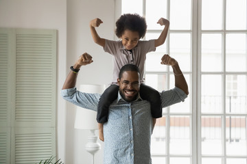 Small son sit on strong dad shoulders showing biceps. African family enjoy activity games at home,...