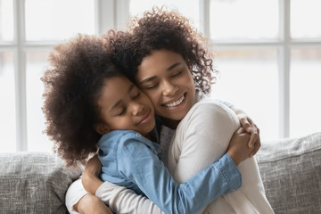 Seated on sofa african loving mother strong cuddles little daughter gives her love and caress show...
