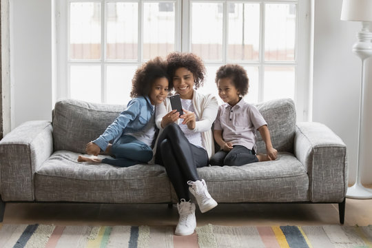 African ethnicity young mother sitting on couch in living room with little kids having fun hold smartphone looking at screen taking selfie photo. Mom teaches small daughter and son using new cool app