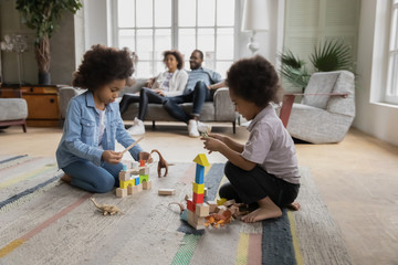 African kids play on floor with wooden colorful bricks set and dinosaur toys while parents rest on...