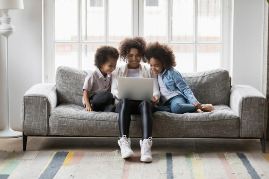 Single African biracial mother and little children sitting on couch using laptop, family enjoy distant talk by videocall, spend time in internet buying, e-commerce users, cartoons or video fun concept