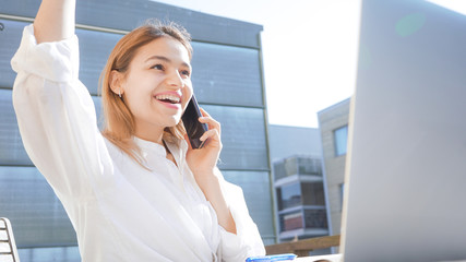 Confident business woman on a phone call. Happy and smiling girl working from home on her balcony on a sunny day. Using computer and phone. 