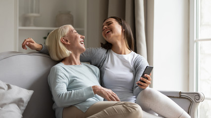 Obraz premium Overjoyed mature mother and millennial grown-up daughter relax on sofa in living room laugh watch funny video on smartphone, happy senior mom and adult girl child have fun using cellphone together