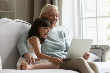 Overjoyed grandmother rest on sofa at home with little granddaughter have fun using laptop together, happy elderly grandparent relax on couch with cute grandchild watch funny videos on computer
