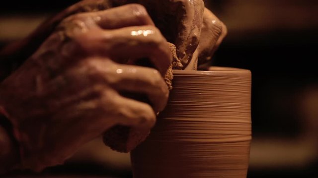 Young handsome sculptor shapes the clay product with pottery tools. Artisan potter prepares material for his pottery. Strong man hands working clay on potter's wheel. Work close-up.