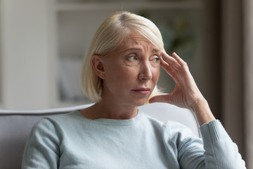 Upset senior woman sit on couch at home look in distance mourning, sad distressed mature female lost in thoughts, miss old days, thinking or pondering, feeling lonely, elderly solitude concept