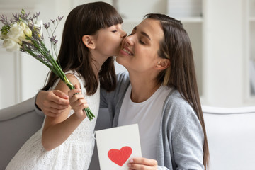 Cute little girl child kiss young mom make birthday surprise present flowers and postcard, loving small preschooler daughter congratulate happy mother with anniversary greeting with bouquet and card