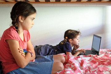 Bored young sister girls using a laptop in bedroom while forced to stay at home because government...