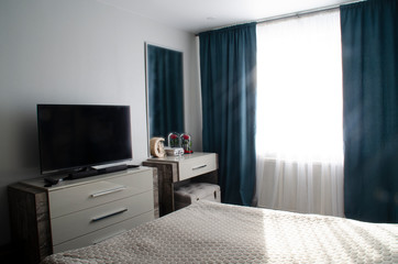 The interior of a modern bedroom in gray and turquoise tones. TV, chest of drawers, dressing table with mirror, bed, curtains.