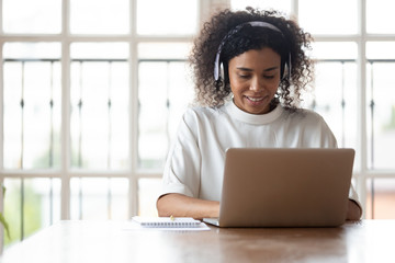 Smiling African ethnicity businesswoman wearing headphones using laptop at work, typing message...