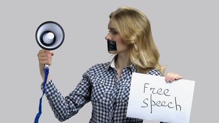 Young woman tries to speak in megaphone with taped mouth. Free speech concept. Grey isolated background.