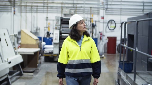 Serious female engineer walking at factory. Front view of concentrated technician walking at plant and looking at working offset printing machine. Industry concept