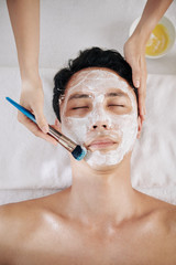 Beautifcian using brush when applying moisturizing mask on face of Asian customer, view from above