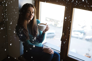 Cute girl sitting with a cup of hot cocoa or coffee by the window in winter with snow background.