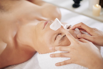 Young man with blackhead remover sticker on his nose enjoying rejuvenating face massage in beauty salon