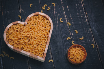 Wooden heart shaped box with pasta or raw macaroni and brown pot on black wooden background, copy space. Carbohydrates diet, healthy eating habits and vegetarian food concept.