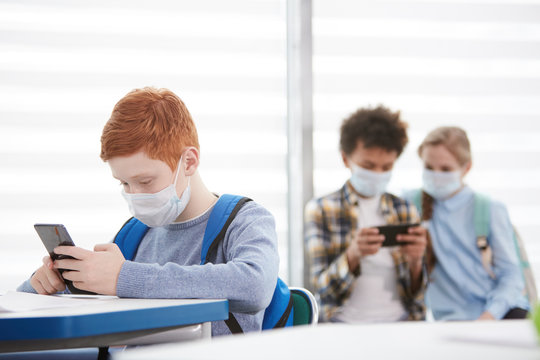 Multi-ethnic Group Of School Children Wearing Face Masks During Epidemic, Focus On Red-haired Teenage Boy Using Smartphone In Foreground, Copy Space