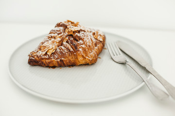 Fresh almond croissant with fork and knife on the plate. Powdered sugar and almond petals. White background. Copy space. 