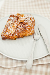 Homemade pastry. Fresh almond croissant on light plate with fork and knife sprinkled with powdered sugar and almond petals. Background of linen towel. 