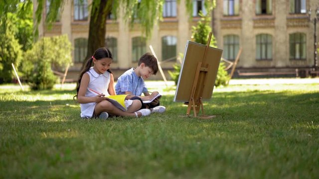 Boy and girl junior school students are sitting outdoors and doing homework. Girl looking thrue her copybook while boy writes in copybook with white board infron of them.