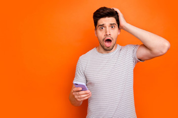 Photo of crazy millennial guy hold telephone open mouth read terrible bad news epic fail website advert hand on head wear striped t-shirt isolated vivid orange color background