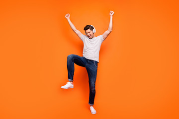 Full size photo of funky cool guy cheerful party mood chilling listening earphones raise hands dancing wear striped t-shirt jeans shoes isolated bright orange color background