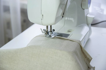 A modern sewing machine on a white table sews from linen fabric.