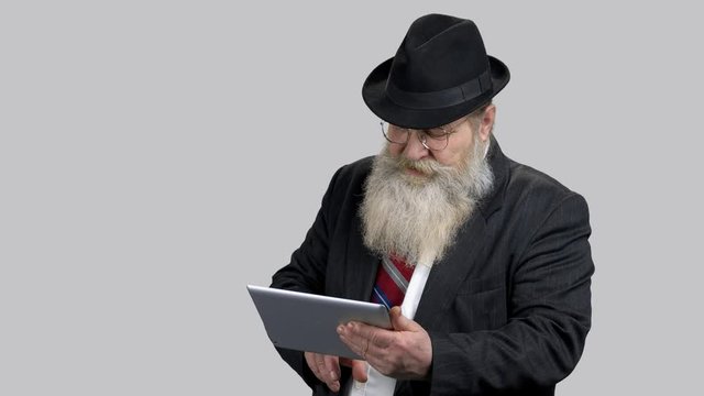 Old businessman swiping and touching tablet pc screen. Portrait of grandpa wearing suit, hat and modern device. Grey isolated background.