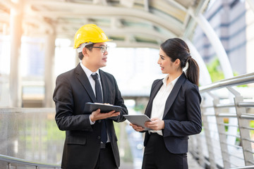 asian businesswoman and a businessman talking to each other planning and brain storming in business project, holding smart tablet device and clipboard, within an urban city district in the background