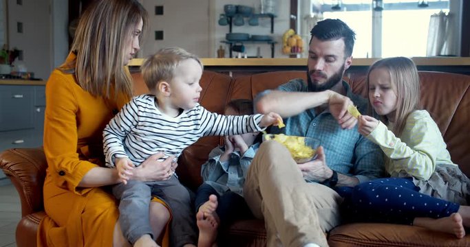 Happy relaxed family time at home, young Caucasian mom, dad and three children sit together eating snacks slow motion.
