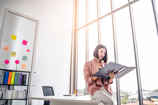 Beautiful Female Asian Worker Holding A Folder File Of Documents And Information On Work Or Business Looking, Reading, Learning From The Documents, While Holding A Pen Working  Office Environment.