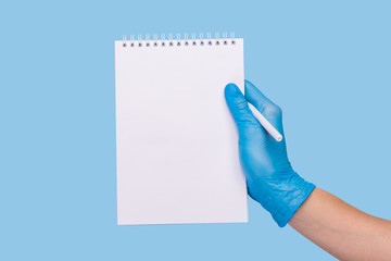 A female hand in a medical glove holds a tablet for writing and a pen on a blue background.