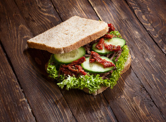 tuna sandwich with green lettuce leaves, dry tomatoes and fresh cucumbers, at rustic table background