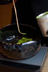 weighing green powder matcha tea in dark ceramic bowl on scales, macro closeup