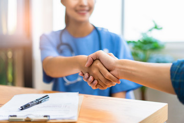 Close up of asian female doctor shaking hands with patient in greeting and respect of doctor helping and support, with document forms and contracts or patients on desk within a hospital office room