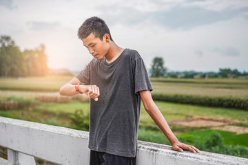 male asian teenager looking at watch checking running distance and heart rate after running and exercising, wearing shirt, short and running shoes, with tree, nature and cloudy sky in the background