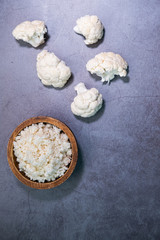 Cauliflower rice on a wooden bowl and cauliflower pieces on concrete background. Vertical picture. Top view.