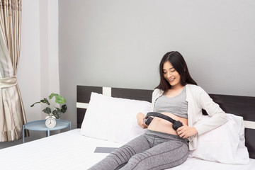 beautiful asian pregnant woman holding and using headphone placing on pregnant belly, letting the baby listening inside, smiling with happiness siting leaning on white wooden bed relaxing in bedroom